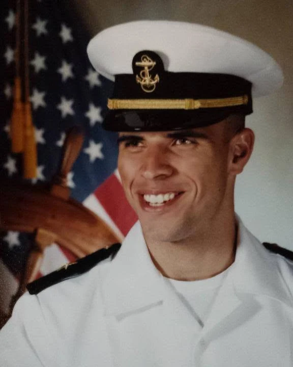 Dominique Ramirez as a Navy Midshipman, in service dress whites, photographed in front of an American flag and a ship's wheel